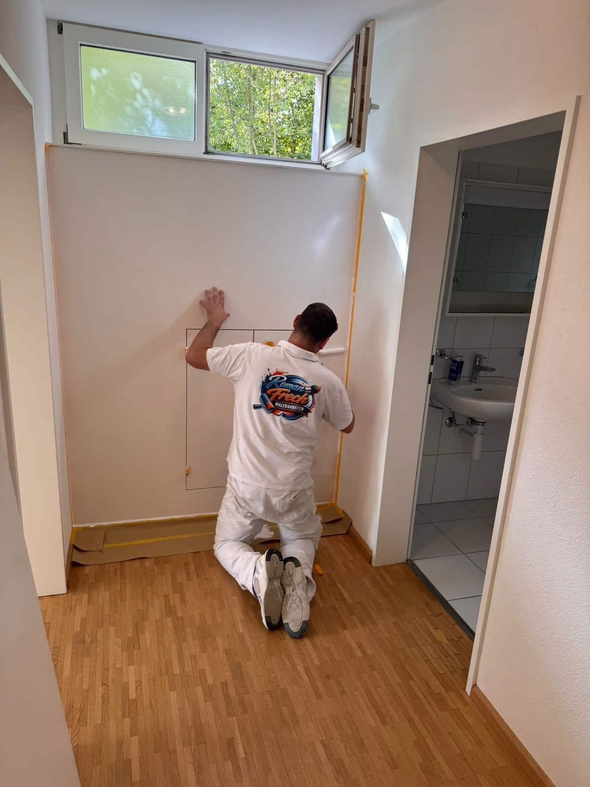 Roman Frech prüft im abgeklebten Flur die Wand von Hand, Logo Roman Frech Malerarbeiten auf dem Shirt