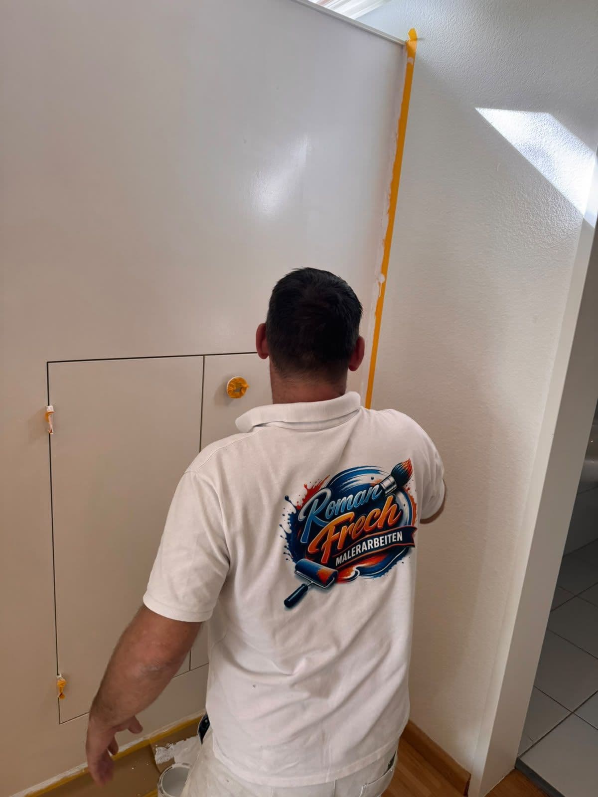 Roman Frech von hinten beim Streichen eines Flurs, Logo Roman Frech Malerarbeiten auf dem Shirt