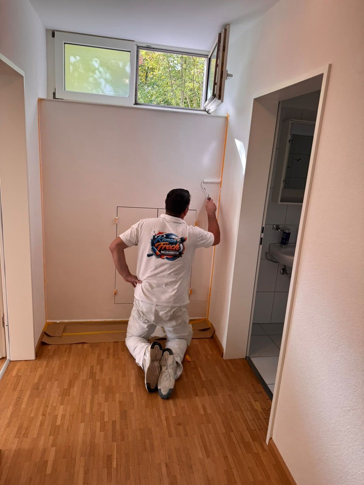 Roman Frech streicht mit der Farbrolle eine Wand im Flur, Logo Roman Frech Malerarbeiten auf dem Rücken